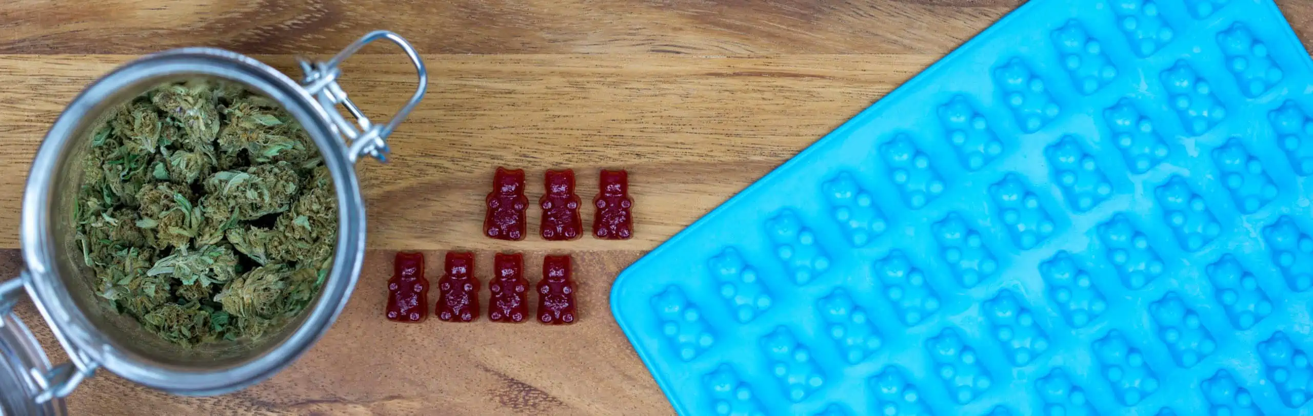 A container of cannabis buds sits next to red RSO gummies and a blue gummy bear mold in preparation for a RSO gummies recipe.