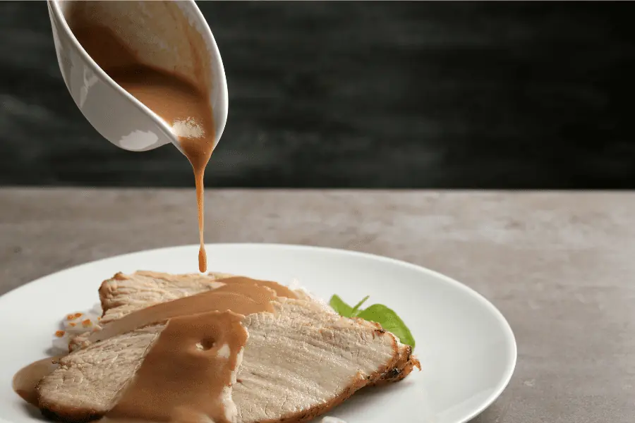 a white gravy container pours brown gravy over a few pieces of turkey on a white ceramic plate.