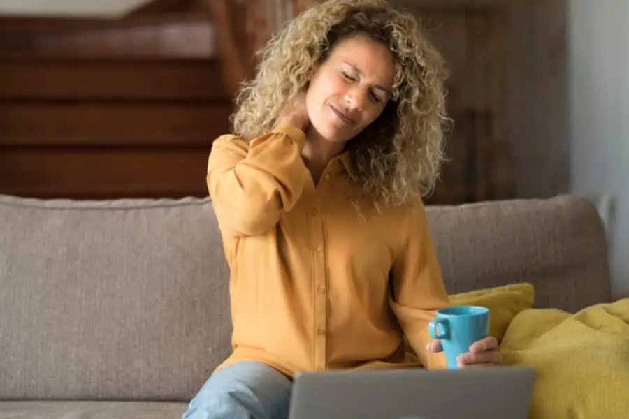 A women sits on a couch rubbing her neck to ease symptoms of stiff person syndrome