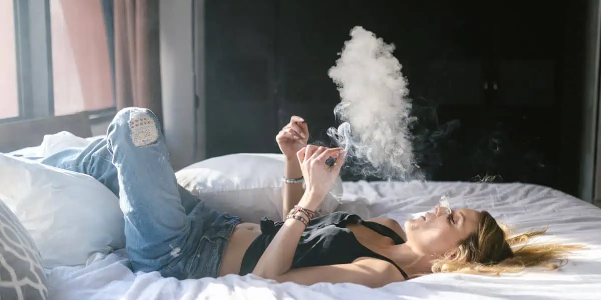 A woman lays on a bed with white sheets smoking a cannabis joint and exhaling smoke into a cloud.