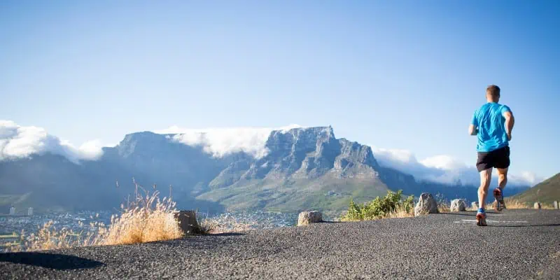 A beautiful light blue sky in the distance can be seen in this photo, along with a range of mountains, while in the up-close focus, there is a man wearing shorts and a blue shirt actively running, depicting the benefits of adaptogens for endurance and weight loss.