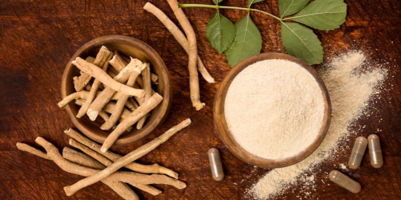 On a wooden rustic table sits a bowl of Ashwagandha in its complete stick-like form, along with a green leaf, a bowl of ground herbs, and capsules beside it.