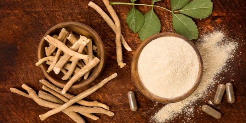 On a wooden rustic table sits a bowl of Ashwagandha in its complete stick-like form, along with a green leaf, a bowl of ground herbs, and capsules beside it.