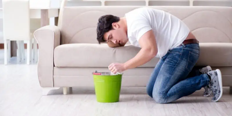This photo shows a gentleman on his knees leaning forward and leaning on a couch, and into a bucket, indicating an adverse effect from cannabis known as cannabis hyperemesis syndrome