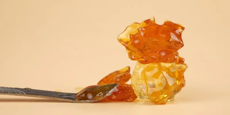 The image features a peach colored backdrop with a glass-like cannabis concentrate known as "shatter" which presents itself as a crystalized version of cannabis that shatters when broken.