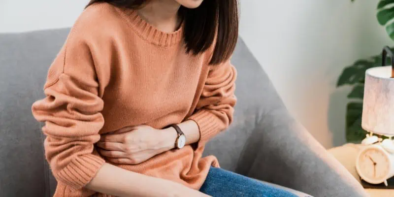 A woman sits on a gray couch and holds her stomach in pain, from the symptoms of crohn's disease.