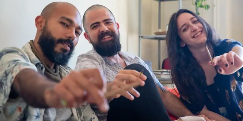 This photo depicts a group of people including two men with bears and a woman, all holding a cannabis joint in there hands as they pass it directly to you, the viewer.