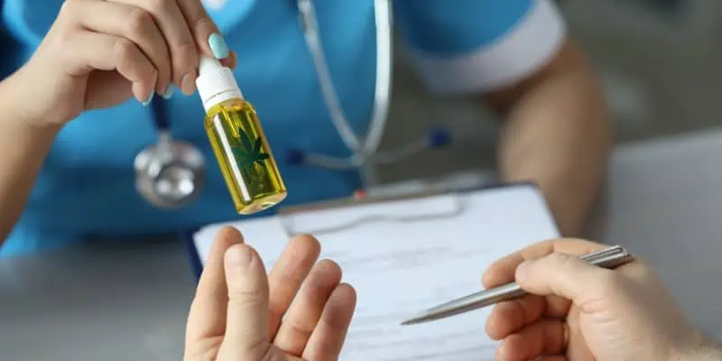 This image depicts a medical professional wearing light blue scrubs and a stethoscope as they hand a vial of cannabis oil to another person with their left hand.