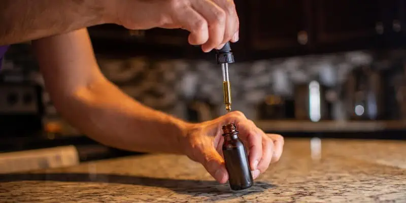 The photo shows a person extracting a cannabis infused oil from a tincture bottle on a granite counter in their kitchen.