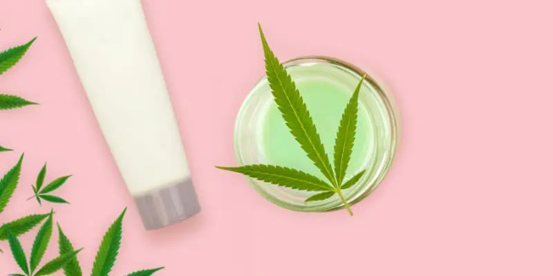 Placed on a bright bubblegum pink background, this image shows a container filled with a pistachio green lotion infused with cannabis and a white lotion tube next to it followed by green marijuana leaves.