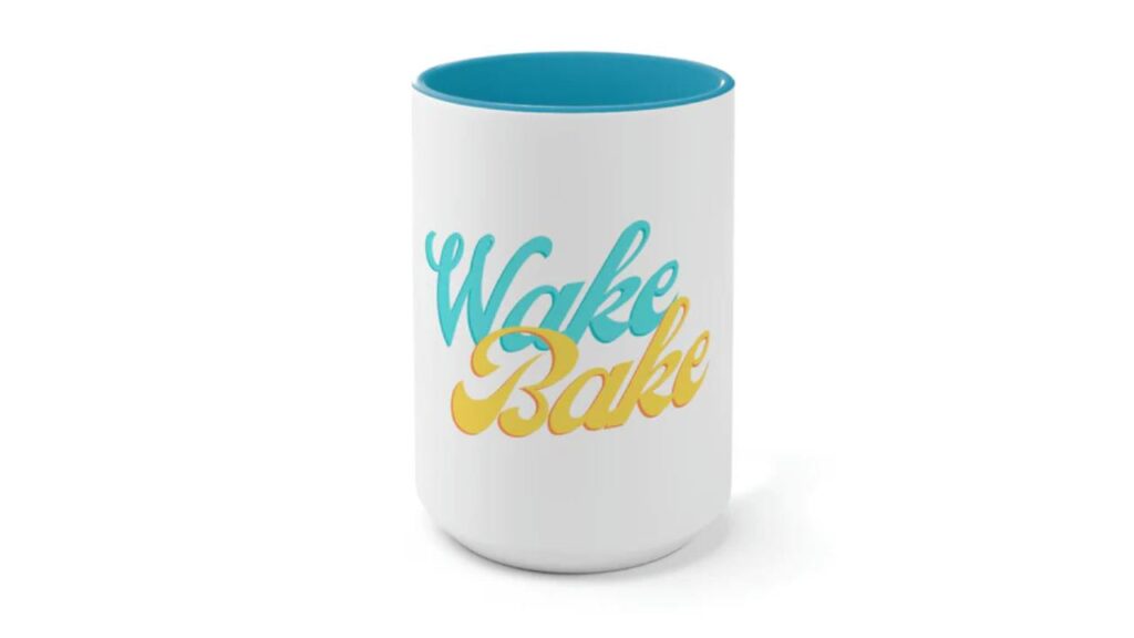 Placed front and center againt a white background, this photo shows a 15 ounce white coffee mug with a two tone blue color on the inside, and reads "wake and bake" on the front in blue and yellow colors.