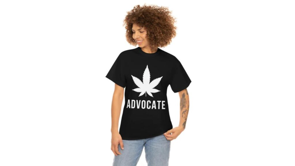 The photo depicts a woman with a tattoo on her left arm, wearing a black t-shirt that reads "advocate" below a white cannabis leaf.