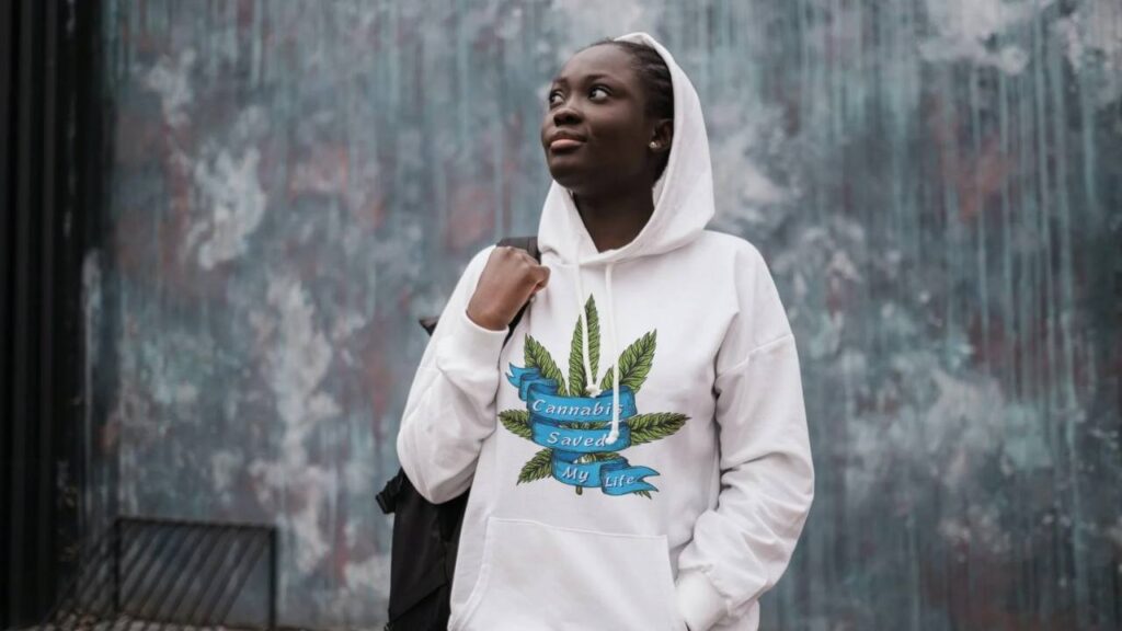 In this photo, a fair-skinned woman in her 20's looks into the distance as she carries her backpack and wears a white cotton blend hoodie with a green cannabis leaf on the front, that features a blue ribbon wrapped around it that reads "cannabis saved my life".