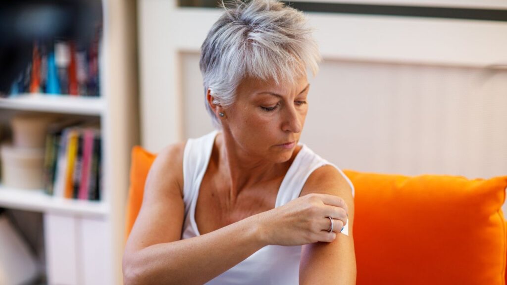 A woman sitting on a couch is depicted as she applies cannabis transdermal patches onto her arm.