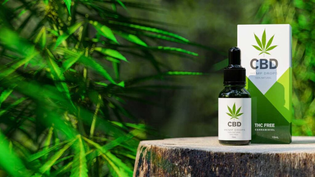 A CBD hemp drops product is shown, placed on top of a tree trunk with a background of hemp leaves growing in the distance.