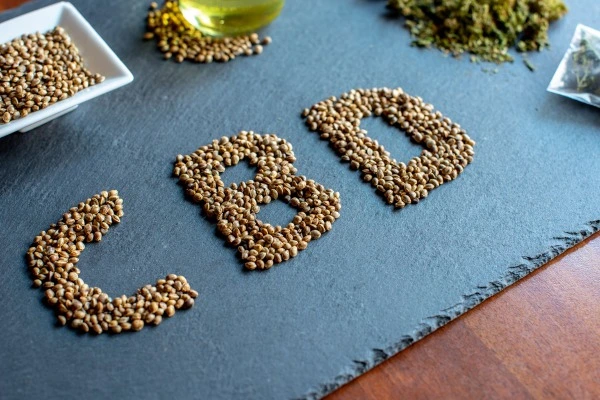 A photo shows tan cannabis seeds arranged on light blue paper to spell out CBD. The paper sits on a brown wood grain desk.