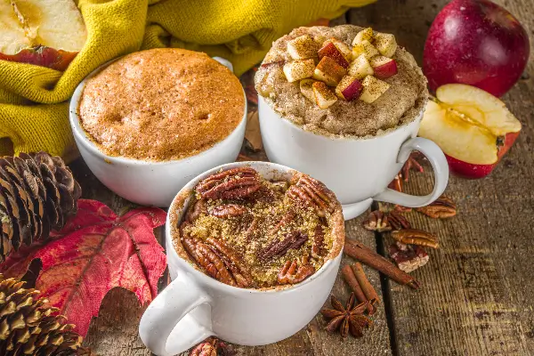 Three mugs of apple spice cannabis infused cakes are displayed on a wood table. One cake has apples and another pecans on top. 