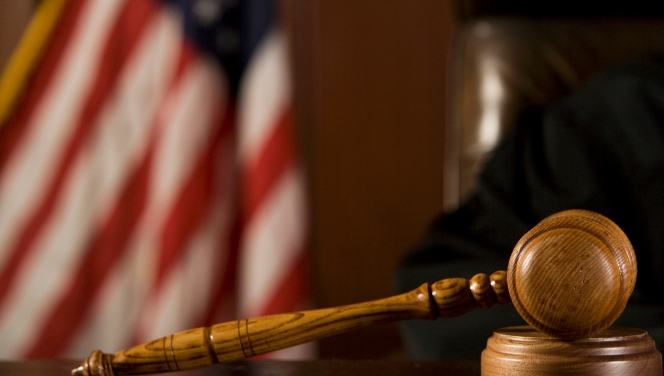 A wooden gavel rests on a desk inside a courtroom with a judge and American flag blurred in the background