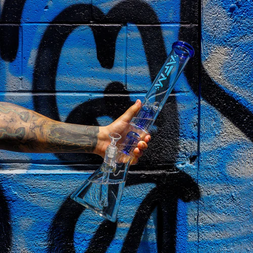 A tattooed hand holds a clear glass bong against a blue graffiti-covered wall.