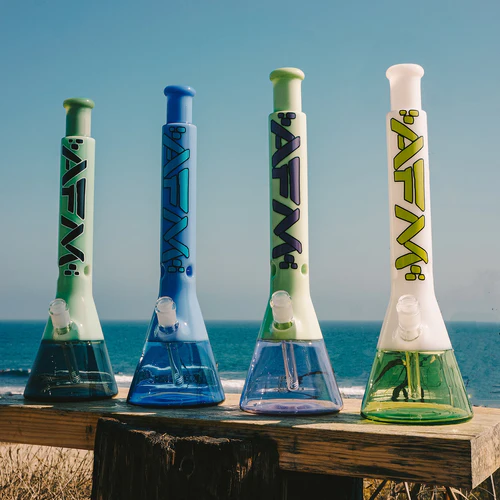 Four colorful bongs with "afm" branding lined up on a wooden surface against a beach background.