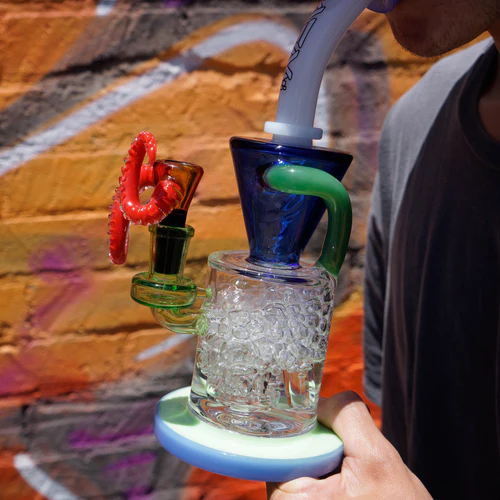A person using a colorful, intricate glass bong outdoors, with a vivid graffiti wall in the background.