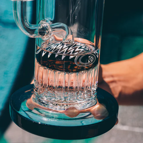 A close-up of a transparent reversal line glass bong filled with water, featuring a spiral percolator, set against a bright, blurred background.