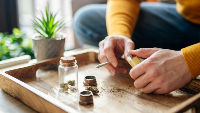 A person in a yellow sweater prepares a cannabis joint on a wooden tray with scattered cannabis, a grinder, and small glass jars.