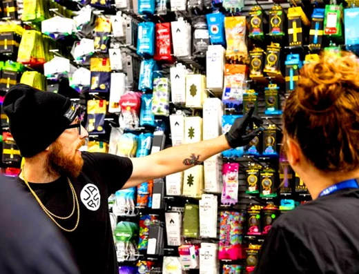 A man in a black beanie and gloves points at a colorful wall display of vaping products while talking to two people, inside what appears to be Jars Cannabis, a cannabis dispensary near Grand Rapids 40th St.