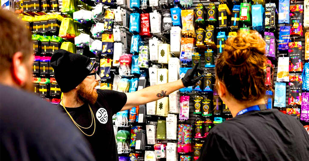 A man in a black beanie and gloves points at a colorful wall display of vaping products while talking to two people, inside what appears to be Jars Cannabis, a cannabis dispensary near Grand Rapids 40th St.