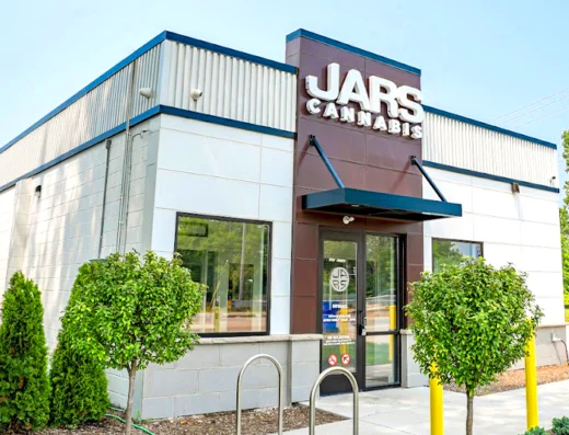 A modern, small white and gray building with blue trim displays the "JARS CANNABIS" sign above its entrance. This inviting cannabis dispensary features glass doors, bike racks, trimmed hedges, young trees, and a backdrop of greenery and clear sky.