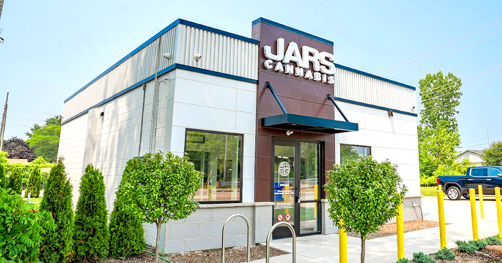 A modern, small white and gray building with blue trim displays the "JARS CANNABIS" sign above its entrance. This inviting cannabis dispensary features glass doors, bike racks, trimmed hedges, young trees, and a backdrop of greenery and clear sky.