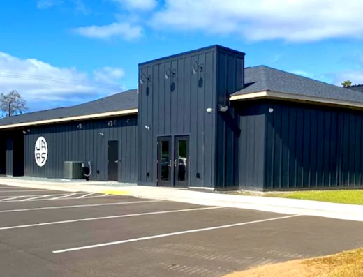 A modern, dark gray, single-story building in Iron River, featuring two large circular JARS Cannabis logos. This cannabis dispensary has a flat and sloped roof, glass double-door entrance, marked accessible parking, grass, and trees in the background.