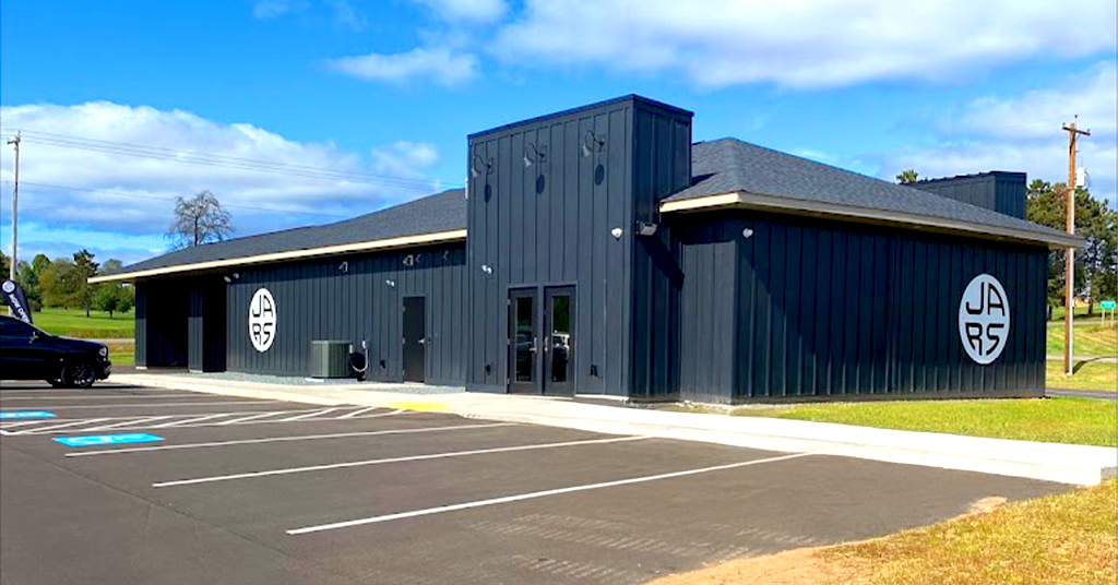 A modern, dark gray, single-story building in Iron River, featuring two large circular JARS Cannabis logos. This cannabis dispensary has a flat and sloped roof, glass double-door entrance, marked accessible parking, grass, and trees in the background.