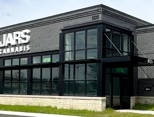 Jars Cannabis Oxford Michigan A modern, gray brick storefront with large windows and a stone base sits in Oxford. Bold white letters read “JARS CANNABIS,” marking it as a cannabis dispensary. Green "OPEN" signs glow in the windows, and grass lines the sidewalk at 1225.