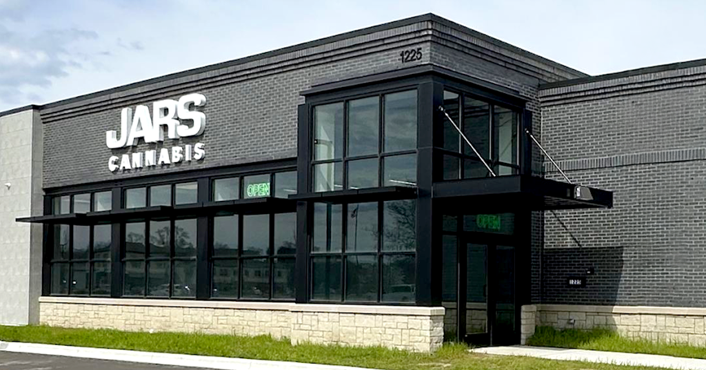 Jars Cannabis Oxford Michigan A modern, gray brick storefront with large windows and a stone base sits in Oxford. Bold white letters read “JARS CANNABIS,” marking it as a cannabis dispensary. Green "OPEN" signs glow in the windows, and grass lines the sidewalk at 1225.