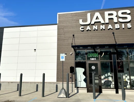 A modern storefront in Muskegon, Michigan, displays a sign reading “JARS Cannabis” above glass doors. A large circular “JARS” logo is on the left wall, with handicapped parking signs, bollards out front, and a neon green "OPEN" sign in the window.