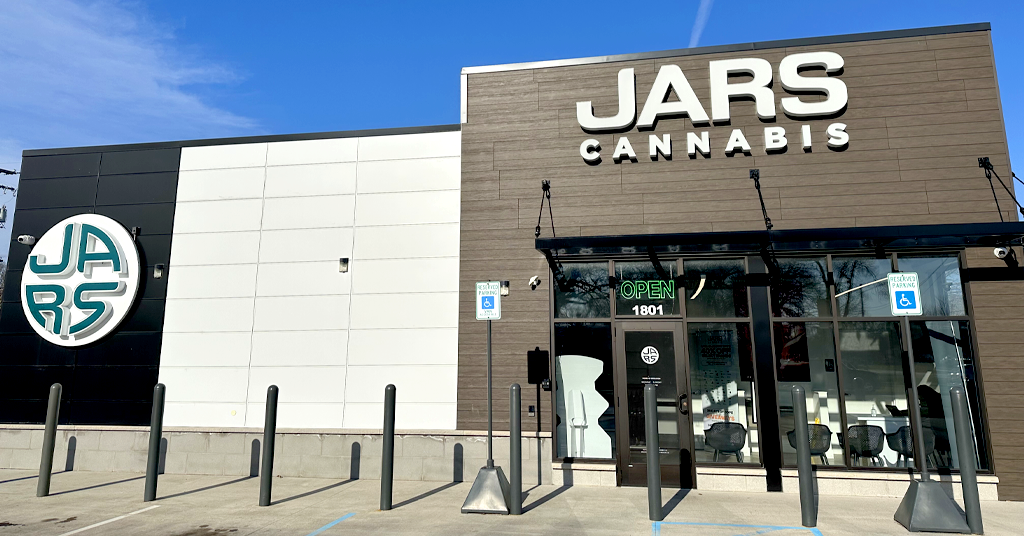A modern storefront in Muskegon, Michigan, displays a sign reading “JARS Cannabis” above glass doors. A large circular “JARS” logo is on the left wall, with handicapped parking signs, bollards out front, and a neon green "OPEN" sign in the window.