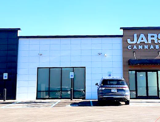 A modern, single-story JARS CANNABIS dispensary in New Buffalo features a white facade with black and wood accents, large front windows, and an awning. Located near the New Buffalo Exit 1, it has a nearly empty lot with multiple handicap spaces and a parked SUV.