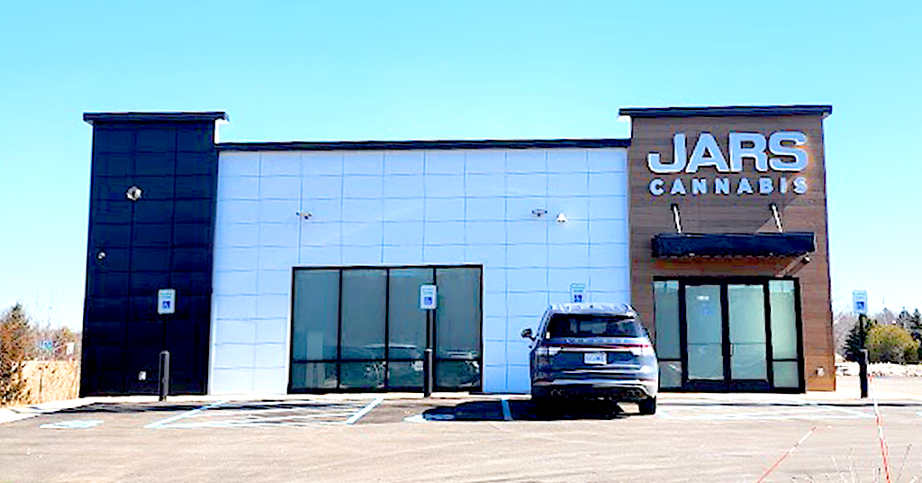 A modern, single-story JARS CANNABIS dispensary in New Buffalo features a white facade with black and wood accents, large front windows, and an awning. Located near the New Buffalo Exit 1, it has a nearly empty lot with multiple handicap spaces and a parked SUV.