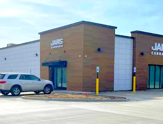 A modern, single-story Cannabis Dispensary with tan and white exterior panels features “JARS CANNABIS” signs above two entrances. Three cars are parked in the lot, yellow bollards line the sidewalk, and trees stand in the background under a clear sky.