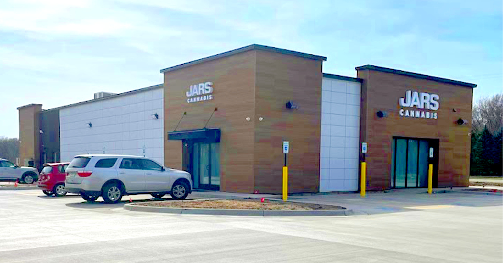 A modern, single-story Cannabis Dispensary with tan and white exterior panels features “JARS CANNABIS” signs above two entrances. Three cars are parked in the lot, yellow bollards line the sidewalk, and trees stand in the background under a clear sky.