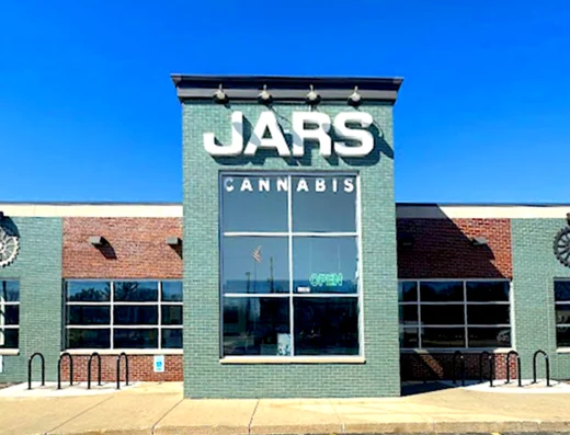 A modern brick building in Oxford Michigan features a central green facade with “JARS CANNABIS” in large white letters. Windows, a bike rack, and a glowing green neon “open” sign complete the inviting scene under a clear blue sky.