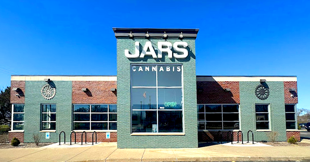 A modern brick building in Oxford Michigan features a central green facade with “JARS CANNABIS” in large white letters. Windows, a bike rack, and a glowing green neon “open” sign complete the inviting scene under a clear blue sky.