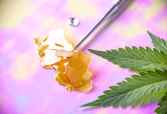 A metal dab tool holds a piece of translucent golden cannabis concentrate on a pink and yellow background. A green cannabis leaf lies beside it, capturing the essence of Urban Farmacy—your source for cannabis delivery in Portland, Oregon.