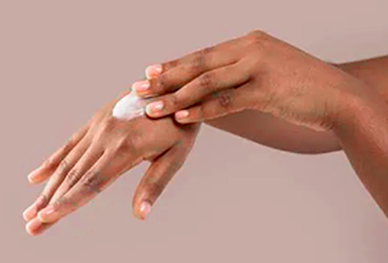 A person with medium brown skin applies a dollop of white lotion to the back of their left hand using their right hand, evoking the self-care culture popular in Portland, Oregon, against a plain light brown background.