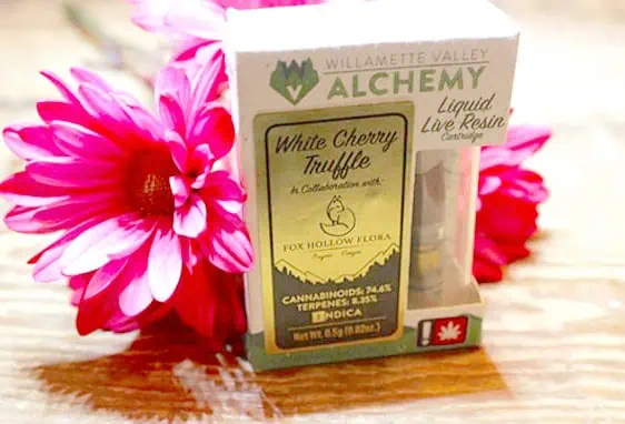 A box labeled “Willamette Valley Alchemy” and “White Cherry Truffle” stands beside bright pink flowers. The Urban Farmacy product, a liquid live resin cartridge (indica, 74.6% cannabinoids), displays a Fox Hollow Flora logo—available via Cannabis Delivery in Portland Oregon.