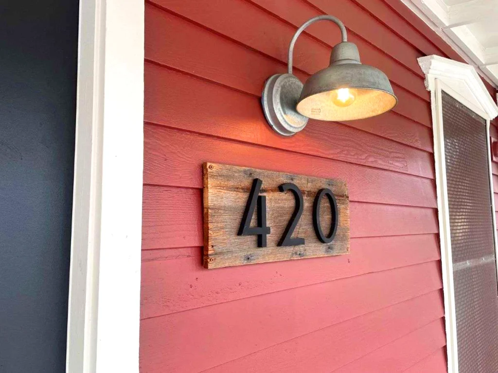 A rustic metal lamp glows above a wooden plaque with black numbers "420" mounted on red siding near a black door and white-framed window, evoking the laid-back spirit of Portland and hinting at nearby cannabis delivery options like Urban Farmacy.