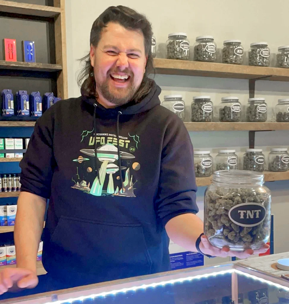 A smiling man with long dark hair and a beard stands behind the counter at Urban Farmacy in Portland, holding a glass jar labeled "TNT" filled with cannabis buds. He wears a black hoodie with a colorful graphic, surrounded by shelves of products and jars.