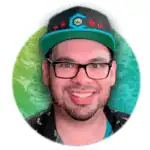 A smiling man with a beard and mustache wears glasses, a black cap with blue and red designs, and a dark shirt. The background is a green and teal textured pattern with a white halo effect around him.