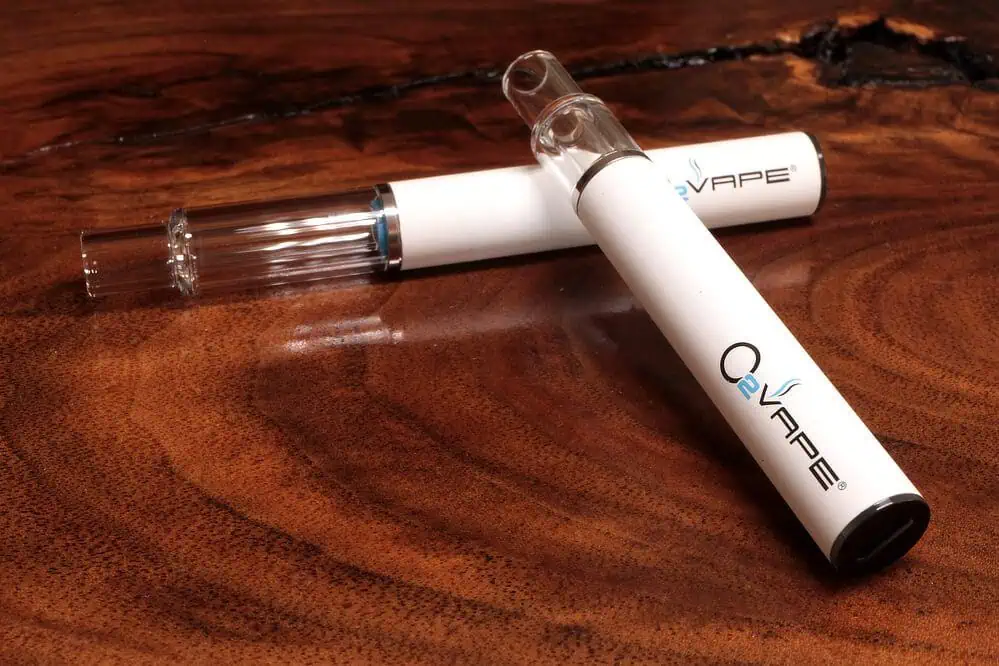 Two white cannabis vape hardware pens with clear glass mouthpieces and "O2VAPE" branding rest on a polished wooden surface. One pen lies flat, while the other is angled across it. The rich wood grain adds texture and warmth to the image.